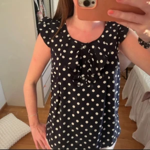 Marinblå prickig blus - Säljer en marinblå blus med vita prickar och volangdetalj vid halsen. Blusen har ärmlös design med små volanger på axlarna och rund halsringning. Perfekt för dig som gillar söta detaljer och mönster.