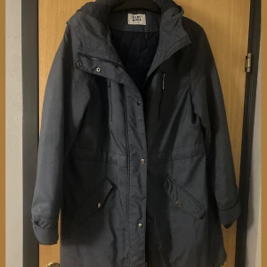 Mörkgrå parkajacka från Vero Moda - Parkajacka i mörkgrått från Vero Moda med quiltat foder och huva. Jackan har praktiska fickor med lock och knappar, samt dragkedja och tryckknappar framtill. Materialet är slitstark polyester och passformen är normal. Perfekt för kyliga dagar.