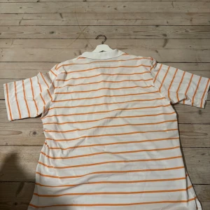Vit och orange randig Polo Ralph Lauren - Vit pikétröja från Polo Ralph Lauren med orangea ränder och klassisk krage. Broderad blå logga på bröstet och två knappar vid halsen. Modellen är Golf Fit och har korta ärmar. Skön bomullskvalitet som passar perfekt till chillade dagar.
