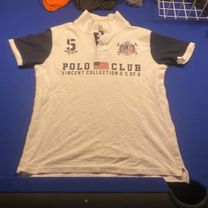 Vit Polo Club henley med tryck - Vit henley från Polo Club Vincent Collection med mörkblå kontrastärmar och tryck på bröstet. Snygga broderade detaljer, amerikansk flagga och klubbemblem. Klassisk krage och knappar framtill. Perfekt för dig som gillar sportig och preppy stil.