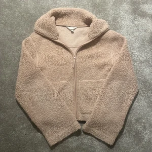 Beige teddyjacka från H&M - Mysig beige teddyjacka med fluffigt material och hög krage. Jackan har en hel dragkedja framtill och långa ärmar, perfekt för kyliga dagar. Enkel och stilren design som passar till många olika outfits.