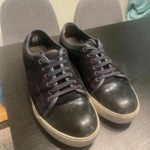 Svarta sneakers från Lanvin mocka - Lanvin sneakers i svart mocka med glansig svart tå och vit platt sula. Klassisk rund tå, snörning och diskret branding på sulan. Snygg och stilren design som passar till jeans eller dressade outfits. Materialmix av mocka och skinn ger en lyxig känsla.