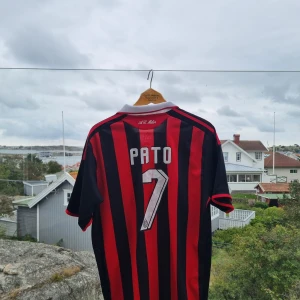AC Milan Pato #7 Adidas fotbollströja - Säljer en klassisk AC Milan fotbollströja med Pato och nummer 7 på ryggen. Tröjan har svart och rött randigt mönster, vit krage och röda detaljer vid ärmslut. Officiell Adidas-logga och klubbmärke på bröstet. Materialet är lätt och andas, perfekt för match eller träning.