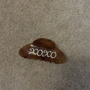 Brun hårklämma med strass XOXO - Snygg brun hårklämma i plast med texten XOXO i glittrande strass. Perfekt accessoar för att piffa upp frisyren och ge en trendig touch till din look.