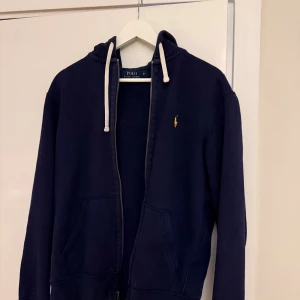 Mörkblå hoodie från Polo Ralph Lauren - Säljer en mörkblå hoodie från Polo Ralph Lauren med vit snörning i huvan och gul broderad logga på bröstet. Tröjan har dragkedja, två fickor fram och är långärmad. Perfekt för chill dagar och enkel att matcha med jeans eller joggers.