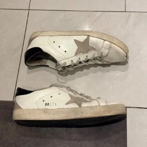 Golden Goose vita sneakers med stjärna - Säljer ett par klassiska Golden Goose sneakers i vitt skinn med grå mockadetaljer och den ikoniska stjärnan på sidan. Skorna har grå snörning, svart hälkappa och en platt sula. Perfekta för dig som gillar en avslappnad och trendig look.