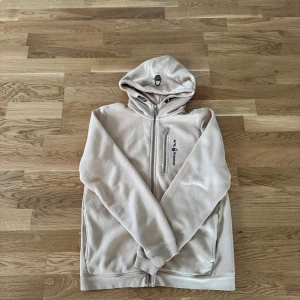 Beige hoodie från Sail Racing - Snygg beige hoodie från Sail Racing med dragkedja framtill och på bröstfickan. Tröjan har huva med snörning, långa ärmar och två sidofickor. Perfekt för chill dagar och har en clean look med diskret logga på bröstet.