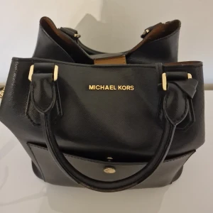 Svart handväska från Michael Kors - Stilren svart handväska från Michael Kors med guldfärgade detaljer och logga framtill. Väskan har både handtag och justerbar axelrem. Har blivit lite lätt böjd då jag har haft böcker i den därav lågt pris. Inköpt för strax under 4000 kr.