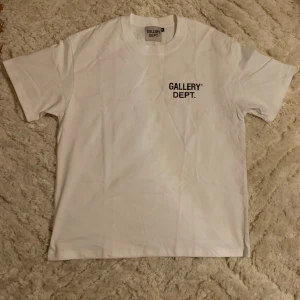 Vit Gallery Dept. t-shirt med tryck - Vit t-shirt från Gallery Dept. med svart tryck på bröstet och stor text på ryggen där det står 'GALLERY DEPT. HOLLYWOOD, CA.'. Klassisk rund hals och korta ärmar. Materialet är mjuk bomull och passformen är normal. Perfekt för dig som gillar streetwear och tydliga loggor.