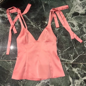 Rosa satinlinne med knytband - Supersöt rosa linnetopp i satin med v-ringning och breda axelband som knyts i rosetter. Toppen har en lätt utsvängd passform och slät yta som ger en lyxig känsla. Perfekt för dig som vill ha en feminin och trendig look.