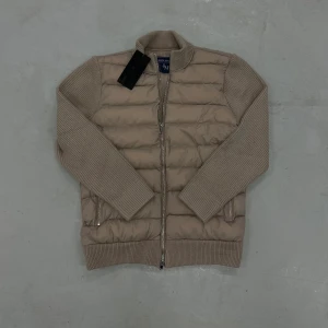 Beige pufferjacka från Leon Marcel - Snygg beige cardigan från Leon Marcel. Jackan har stickade ärmar och rygg i matchande beige ton, quiltad framsida och tvåvägsdragkedja. Perfekt för dig som gillar en clean look med coola detaljer och vill ha något som sticker ut.