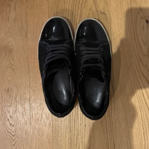 Svarta sneakers från Valentino - Snygga svarta sneakers från Valentino med blank tå och sidor i mocka. Skorna har rund tå, snörning och vit platt sula som ger en clean kontrast. Perfekta för dig som gillar stilrena och exklusiva märken. Insidan är i svart skinn för extra komfort.