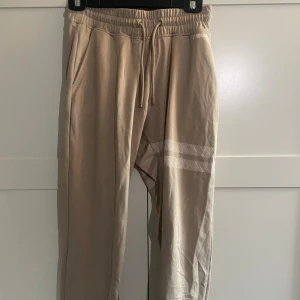 Beige mjukisbyxor från Stronger - Snygga beige mjukisbyxor från Stronger med elastisk midja och snörning. Byxorna har två sidofickor och vita ränder med prickigt mönster på ena benet. Mjukt material och sköna att träna i! Finns matchande tröja.