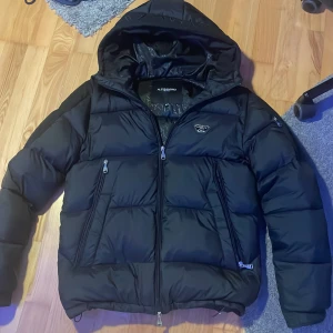 Svart pufferjacka Alessandro Zavetti - Svart pufferjacka från Alessandro Zavetti med huva och silverfärgade dragkedjor. Jackan har quiltad design, två sidofickor med dragkedja och en snygg metallbadge på bröstet. Perfekt för kalla dagar och har en modern, urban look.