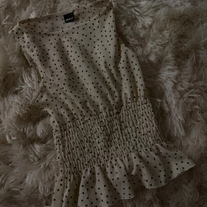 Prickig ärmlös topp från Gina Tricot - Supersöt beige ärmlös topp från Gina Tricot med svarta prickar. Toppen har smockad midja och volangkant nertill för en extra feminin touch. Perfekt att styla med jeans eller kjol för en trendig look.