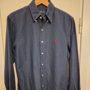 Mörkblå skjorta från Ralph Lauren - Snygg mörkblå skjorta från Ralph Lauren i slim fit med klassisk krage och långa ärmar. Skjortan har knappar framtill och en röd broderad Polo-logo på bröstet. Tillverkad i mjuk bomull som känns skön mot huden. Perfekt för dig som gillar stilrena plagg med ikonisk touch.