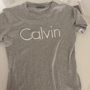 Grå t-shirt från Calvin Klein Jeans - Snygg grå t-shirt från Calvin Klein Jeans i mjuk bomull. Framsidan har ett stort broderat 'Calvin'-tryck i vitt. Modellen är klassisk med rund hals och korta ärmar, perfekt för en chill och stilren look.