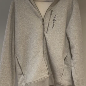 Grå hoodie från Sail Racing med dragkedja - Snygg ljusgrå hoodie från Sail Racing med hel dragkedja framtill.  Tröjan har huva med snören för spänning och snygg detalj. Perfekt för chill dagar eller när du vill ha en clean look. Tröjan är knappt använd då den är för stor för mig.