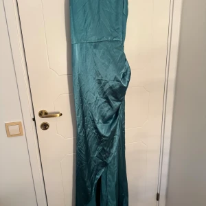 Turkos långklänning från SHEIN - Säljer en turkos långklänning från SHEIN i glansigt material. Klänningen har smala axelband, slits framtill och en snygg knytning i ryggen. Perfekt för dig som vill sticka ut med en elegant och trendig look.