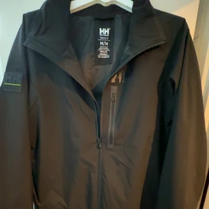 Svart vindjacka från Helly Hansen M - Svart vindjacka från Helly Hansen med Helly Tech Performance-tyg. Jackan har dragkedja framtill, en bröstficka med dragkedja och diskret logga på kragen. Regular fit och långärmad, perfekt för blåsiga dagar. Snygg och stilren design med patch på ärmen. Jag har använt jackan 1 gång men den är för stor. Inga defekter och den är mer eller mindre ny. Skriv vid funderingar .🤗