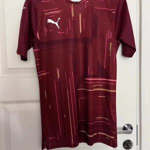 Vinröd fotbollströja från Puma S - Snygg vinröd fotbollströja från Puma med korta ärmar och ett grafiskt mönster i rosa och gult. Tröjan har en vit Puma-logga på bröstet och är tillverkad i ett lätt, ventilerande syntetmaterial som passar perfekt för träning eller match.