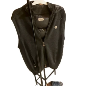 Svart dunväst från Moncler med huva - Svart dunväst från Moncler med stickade sidor och quiltad framsida och rygg. Västen har huva, två dragkedjefickor och Moncler-logga på bröstet. Perfekt för lager-på-lager och snygg till streetwear.