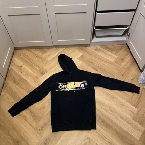 Svart Off-White hoodie med tryck - Svart hoodie från Off-White i storlek S med fet vit och gul grafisk logga på både bröstet och ryggen. Tröjan har huva med snörning och en stor magficka. Mjuk insida och ribbade muddar vid ärmslut och nederkant. Perfekt för dig som gillar streetwear.