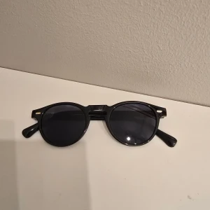 Svarta runda solglasögon - Snygga svarta solglasögon med rund form och klassisk design. Bågarna är i plast och har små silvriga detaljer vid hörnen. Perfekta för att ge din stil en cool touch.