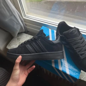 Svarta Adidas Campus sneakers - Säljer ett par svarta Adidas Campus sneakers med klassiska tre ränder på sidan. Skorna har rund tå, platt sula och snörning. Ovandelen är i mocka och insidan är vadderad för extra komfort. Perfekt för dig som gillar stilrena och tidlösa sneakers.