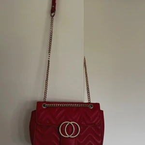 Röd quiltad axelväska med kedja - Snygg röd axelväska med quiltad design och guldfärgad kedjerem. Väskan har ett dekorativt spänne framtill med två ringar och ett elegant mönster i materialet. Perfekt för dig som vill sticka ut med en trendig accessoar.