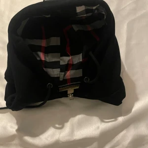 Svart Burberry hoodie med dragkedja - Svart hoodie från Burberry med klassiskt rutigt foder i huvan och insidan. Tröjan har hel dragkedja, snörning i huvan och ribbade muddar. Ikonisk Burberry-logga på bröstet och snygga detaljer i silver på snörena.