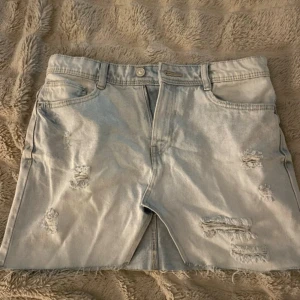 Ljusblå slitna jeanskjol - Säljer ett par ljusblå jeanskjol med slitna detaljer och råa kanter. Klassisk femficksmodell med knapp och dragkedja framtill. Perfekta för en avslappnad och trendig look i sommar. Liten defekt som man kan se på andra bilden men inte som man tänker på 