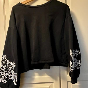 Svart croppad tröja från Zara XL - Svart croppad tröja från Zara i storlek XL med långärmade, ballong-ärmar. Ärmarna har vita broderade detaljer med ett abstrakt mönster. Tröjan har rund hals och är tillverkad i mjuk bomull. 