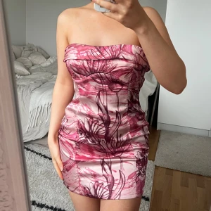 Bershka Klänning 🩷 - Säljer en axelbandslös miniklänning med mönster i rosa och vinröda toner. Klänningen har snörning i ryggen för en snygg passform och är figurnära. Materialet är glansigt och känns lyxigt mot huden. Perfekt för dig som vill sticka ut med en unik look. Från Bershka, storlek S, fint skick 🩷