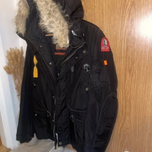 Svart Parajumpers parkajacka med päls - Svart parkajacka från Parajumpers med stor huva och beige fuskpäls. Jackan har flera fickor, gul detaljrem och patch på ärmen. Dragkedja framtill och långa ärmar. Perfekt för kalla dagar med sin rejäla look och coola detaljer.