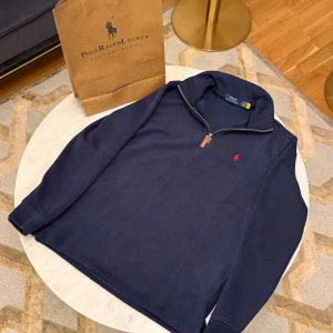 Marinblå half zip tröja Polo Ralph Lauren - Snygg marinblå långärmad tröja från Polo Ralph Lauren med half zip och läderdetalj på dragkedjan. Klassisk röd broderad logga på bröstet. Mjuk bomullskvalitet och normal passform, perfekt för chill dagar.