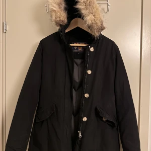 Svart parkajacka Woolrich XS - Svart parkajacka från Woolrich med päls på huvan, dragkedja och knappar framtill. Jackan har två stora fickor med lock och klassisk längre passform. Perfekt för kalla dagar med snygg och tidlös look. OBS: Jackan har ett litet slitage på ärmen (se sista bilden)