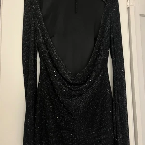 Svart glittrig klänning med öppen rygg - Supersnygg svart klänning med glittriga detaljer och lång ärm. Klänningen har en djup öppen rygg med draperad effekt som ger en extra cool vibe. Perfekt för dig som vill sticka ut med lite extra glitter. Sitter tight och har en kortare längd.