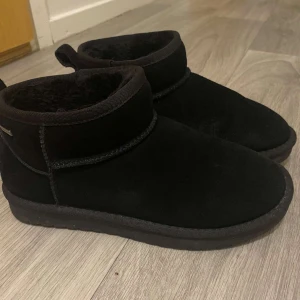 Svarta uggs från Duffy i mocka - Svarta uggs från Duffy med rund tå och platt sula. Skorna är tillverkade i mjuk mocka och har en fluffig insida som håller fötterna varma. Perfekta för kyliga dagar och har en enkel, stilren design med dragflik bak.