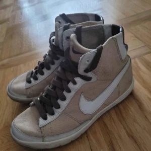 Beige Nike high sneakers strl 35.5 - Snygga beige Nike sneakers med högt skaft och vita detaljer. Skorna har bruna snören, vit swoosh och robust platt sula. Materialet är en mix av textil och skinn, vilket ger en cool kontrast. Perfekt för dig som gillar klassisk streetstil och vill sticka ut med retrovibe.