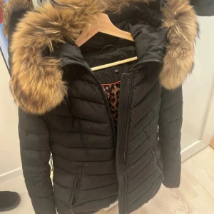 Svart dunjacka med päls från Hollies - Svart dunjacka från Hollies med fluffig päls på huvan och leopardmönstrat foder. Jackan har dragkedja framtill, två sidofickor med dragkedja och quiltad design. Perfekt för kalla dagar och har en snygg, modern look.