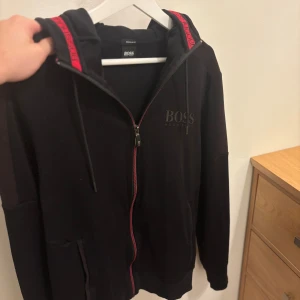 Hugo Boss tröja  - Svart hoodie från Hugo Boss med röd detalj på huvan och dragkedja framtill. Tröjan har huva med snörning, fickor på sidorna och diskret Boss-logga på bröstet. Perfekt för en chill och stilren look.                       Storleken är S/M 