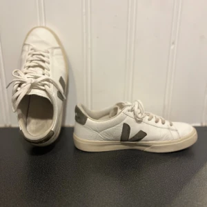 Vita Veja sneakers - Snygga vita sneakers från Veja med gröna mockadetaljer på sidorna och hälen. Skorna har klassisk snörning, platt sula och rund tå. Tillverkade i skinn med Veja-logga på sidan och bak. Perfekta för dig som gillar stilrena och trendiga sneakers. Sitter skönt och inte använda allt för mycket, storlek 41!
