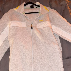 Grå vindjacka från BOSS med dragkedja - Snygg grå vindjacka från BOSS med vita detaljer på bröstet och längs ärmarna, samt gul och grön accent vid kragen. Jackan har hel dragkedja framtill, ståkrage och två sidofickor. Perfekt för dig som gillar sportig och clean stil.