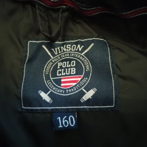 Svart pufferjacka från Vinson Polo Club - Svart pufferjacka från Vinson Polo Club med fuskpäls på huvan och två snedställda fickor med dragkedja. Jackan har quiltad design och detaljer i silverfärg. Perfekt för kyliga dagar.