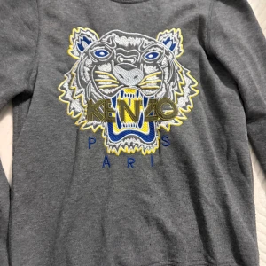 Grå Kenzo sweatshirt med tigertryck - Säljer en grå sweatshirt från Kenzo med ikoniskt broderat tigerhuvud i blått, gult och vitt. Tröjan har rund halsringning och långa ärmar. Perfekt statement-plagg för dig som gillar streetstyle och vill sticka ut.
