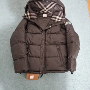 Svart pufferjacka från Burberry - Svart pufferjacka från Burberry med klassisk logotyp på ärmen och rutigt foder i huvan. Jackan har dragkedja och knappar framtill, samt en praktisk ficka med dragkedja. Perfekt för kalla dagar med sin tjocka och quiltade design.