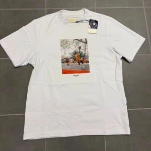 Vit t-shirt Aimé Leon Dore basketmotiv - Vit t-shirt från Aimé Leon Dore med tryck av basketspelare på bröstet. Klassisk passform med rund hals och korta ärmar. Materialet är mjuk bomull och motivet har inslag av orange och svart. Perfekt för dig som gillar streetwear och sportiga vibes.