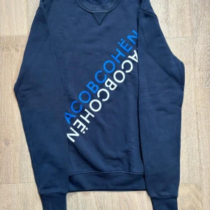 Mörkblå sweatshirt från Jacob Cohën - Snygg mörkblå sweatshirt från Jacob Cohën med diagonal logotyp i blått och vitt på framsidan. Tröjan har rund hals, långa ärmar och ribbade muddar vid ärmslut och nederkant. Perfekt för dig som gillar stilrena och coola prints. Storlek Large
