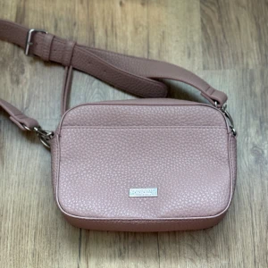 Rosa axelväska från Don Donna - Snygg rosa axelväska från Don Donna med justerbar rem och silverfärgad dragkedja. Väskan är tillverkad i syntetmaterial med en pebble-textur och har en stilren, rektangulär form. Perfekt för att bära dina essentials och ger en cool touch till din outfit.